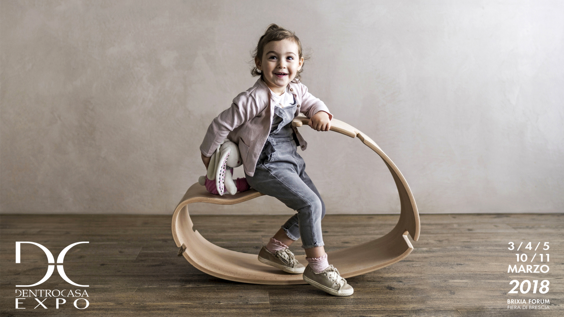 Locandina dell'esposizione del prodotto YaWood di Argo4 presso la fiera DentroCasa Expo a Brescia. Una bambina di un anno e mezzo sorride felice seduta sul cavallo a dondolo in legno curvato Yawood.